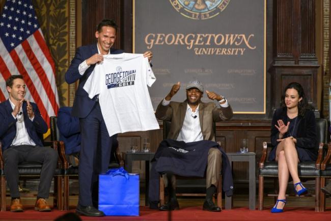 Marc Howard holding a Georgetown Golf shirt on stage with Valentino Dixon